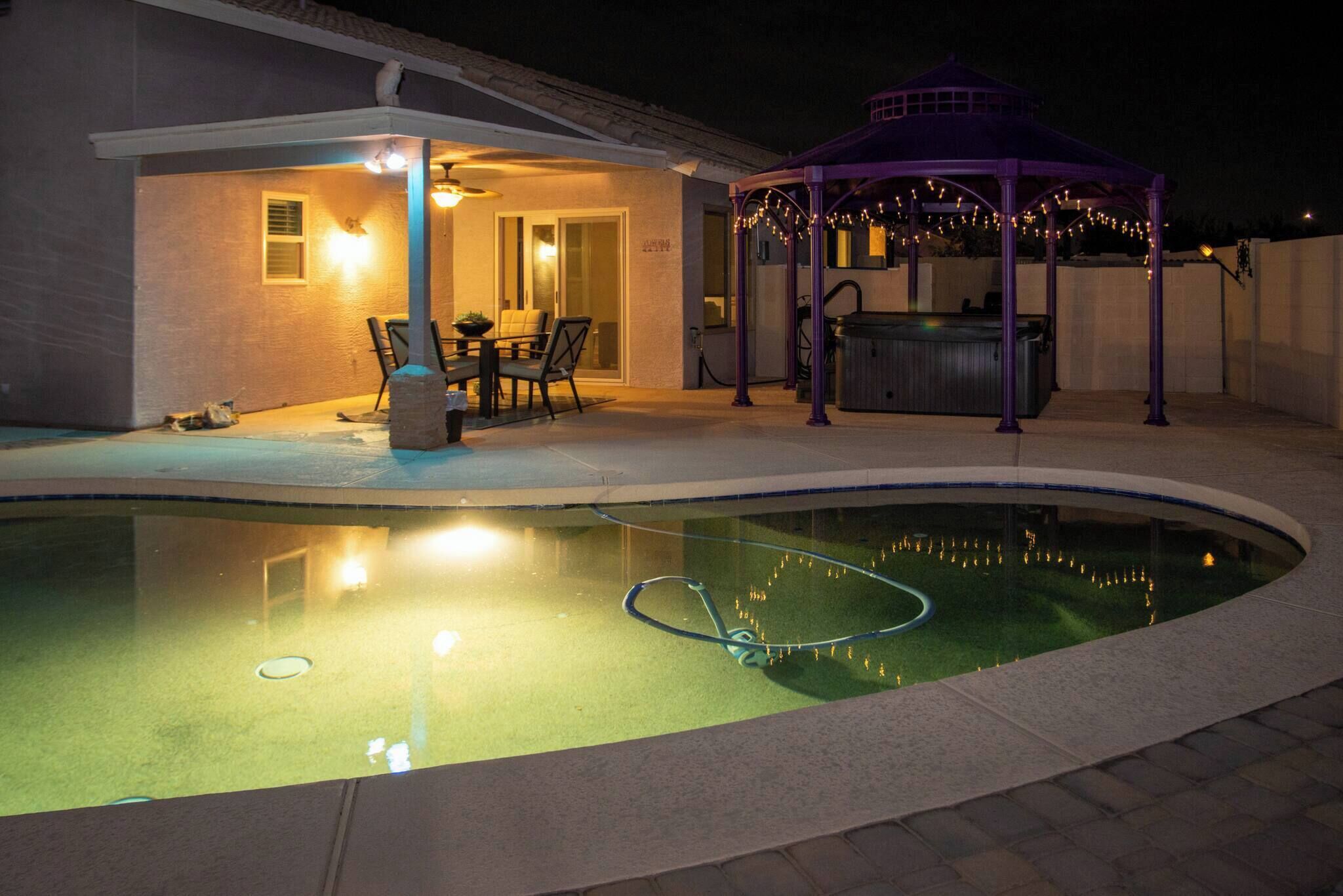 A swimming pool with a hot tub, a covered patio with chairs, and a gazebo.