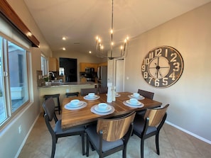 A dining area with a wooden table, chairs, a large wall clock, and a kitchen in the background.