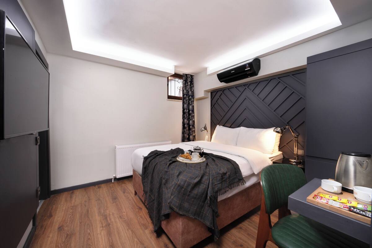 A hotel room with a bed, a desk with a chair, a radiator, a window with curtains, and a ceiling light.