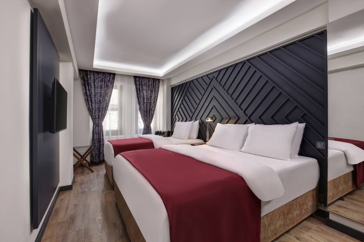 A modern hotel room with two beds, a flat-screen TV, a decorative wall panel, and a window with curtains.