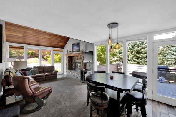 Amplia sala de estar con chimenea, comedor y ventanas grandes con vistas al exterior.