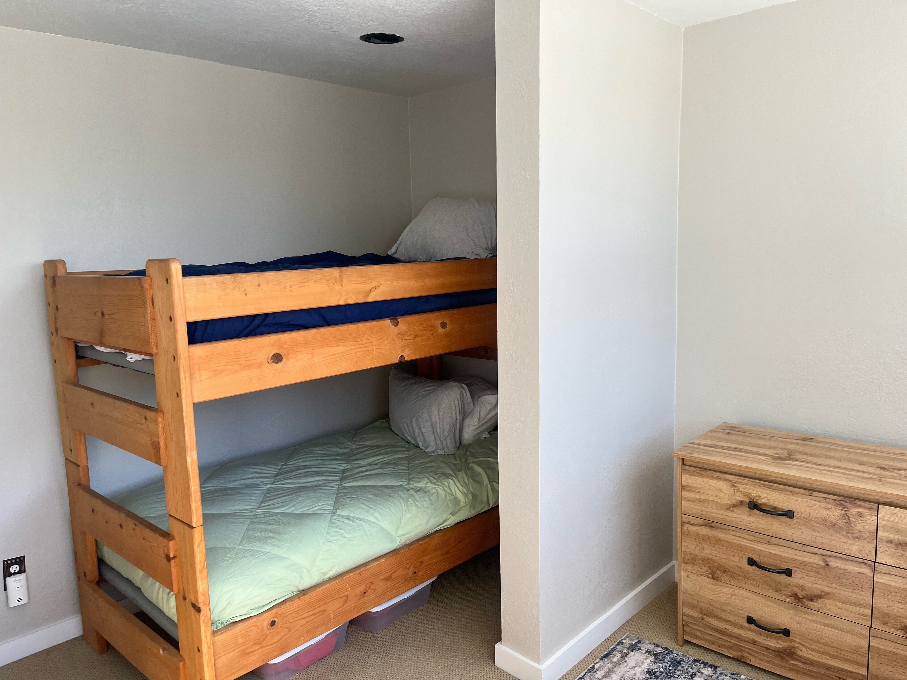 Ein Etagenbett mit Holzbalken, die untere Etage mit einer grünen Bettdecke und die obere Etage mit einer blauen Decke. Dazu ein Holzschrank mit Schubladen zur Rechten.