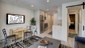 Uma cozinha moderna com eletrodomésticos de aço inoxidável, uma área de refeições com mesa e cadeiras de madeira, e uma sala de estar com sofá e TV exibindo 'STAY LIVING THE DREAM'.
