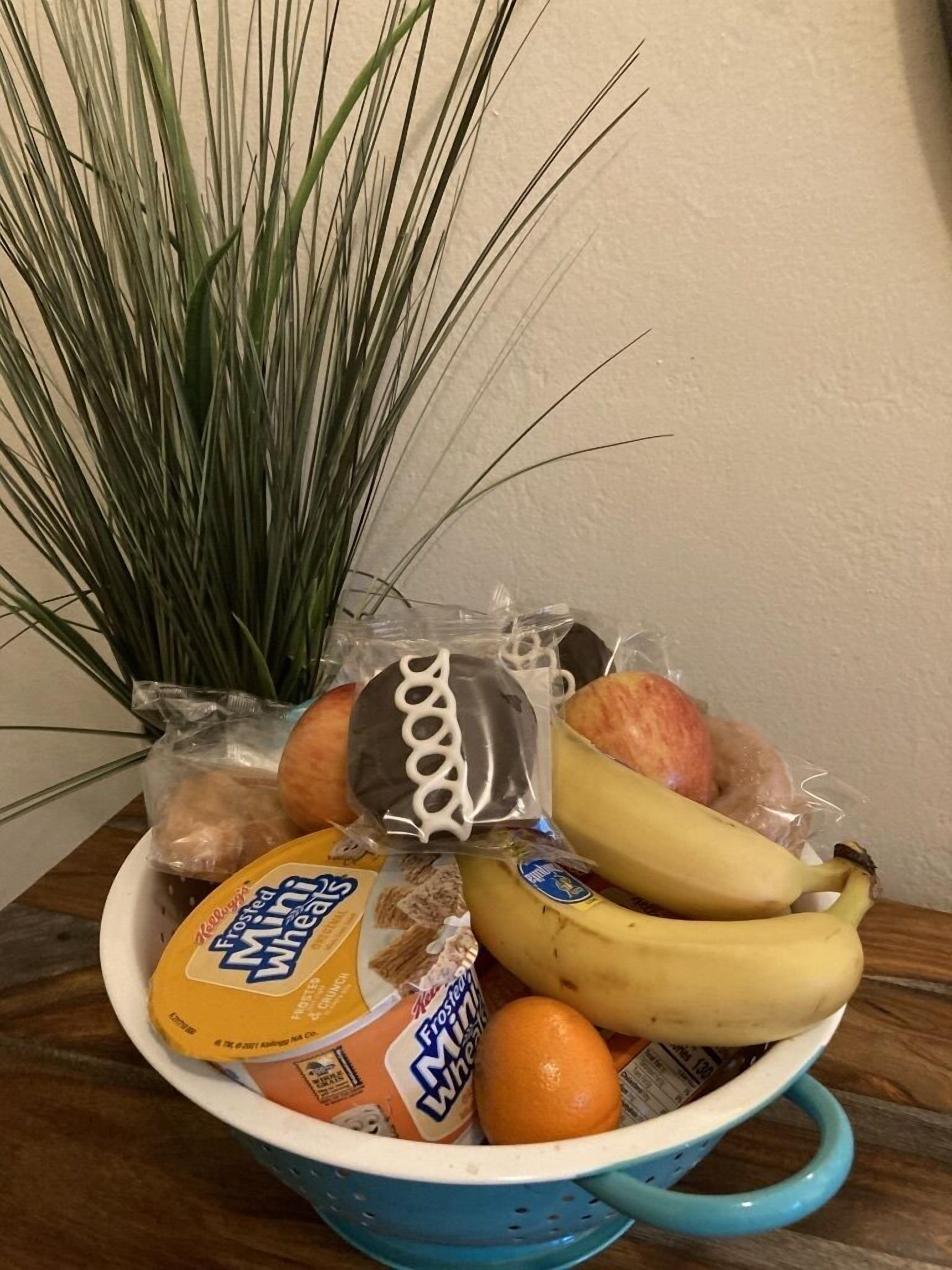 A blue colander containing a variety of food items including a banana, an orange, a package of Frosted Mini Wheats, a chocolate-covered cookie, and a bag of bread.