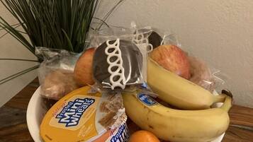 A blue colander containing a variety of food items including a banana, an orange, a package of Frosted Mini Wheats, a chocolate-covered cookie, and a bag of bread.