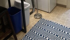 Une cuisine de taille réduite avec un réfrigérateur blanc, une poubelle bleue, un tapis à motifs et une table avec une lampe.