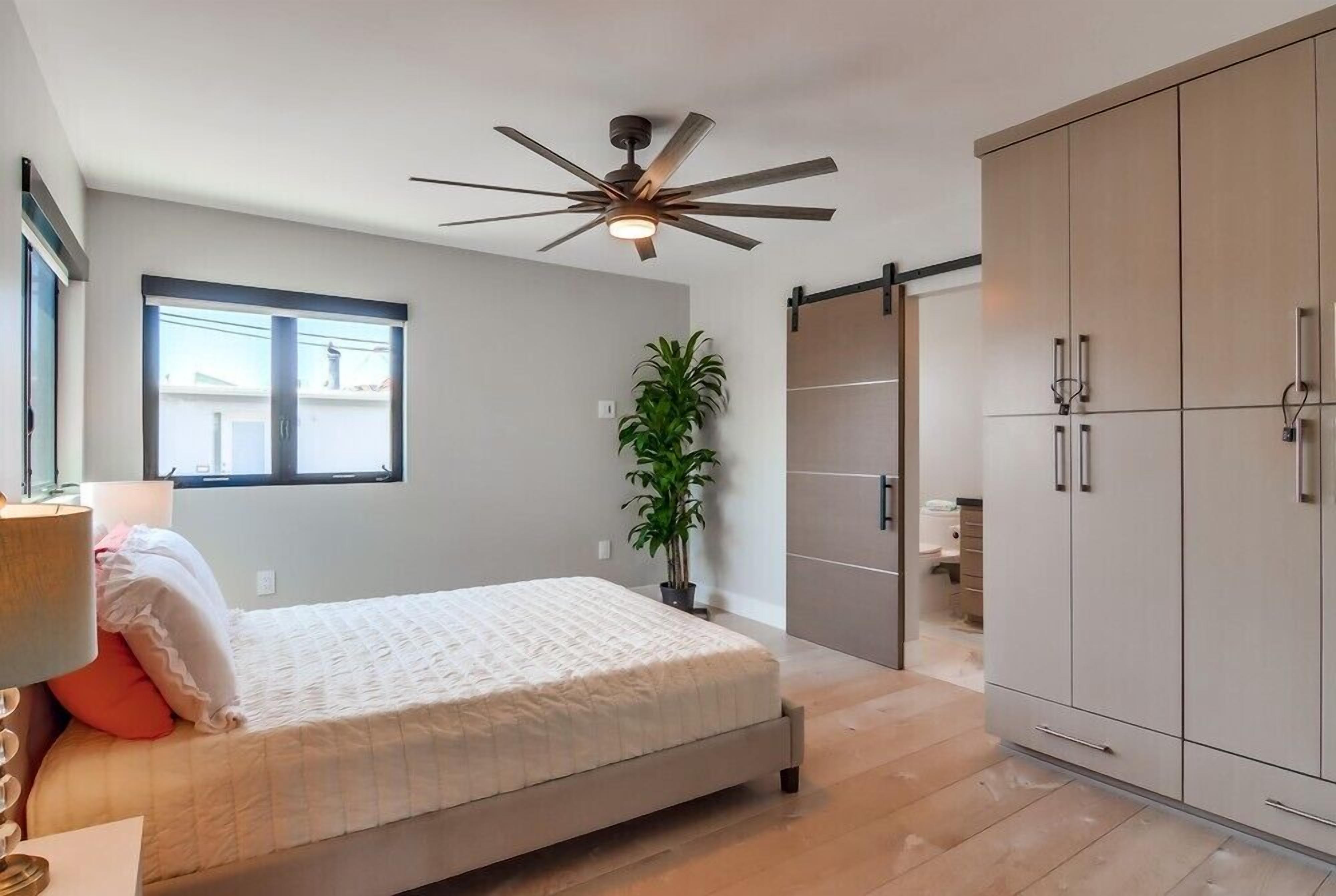 Un dormitorio con cama, ventilador de techo, ventana, planta y armario.
