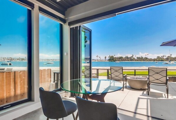 A modern outdoor seating area with a glass table and chairs, overlooking a beach and marina.
