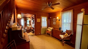 A cozy wooden cabin interior with a piano, a dining area, a living room with a sofa and a chair, and a ceiling fan. - Historic cottage just minutes from downtown Harbor Springs! (Harbor Springs)