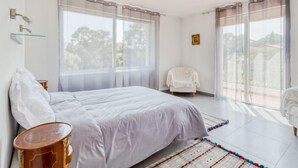 Ein Schlafzimmer mit Bett, Sessel, kleinem Tisch und einem Fenster mit Vorhängen.