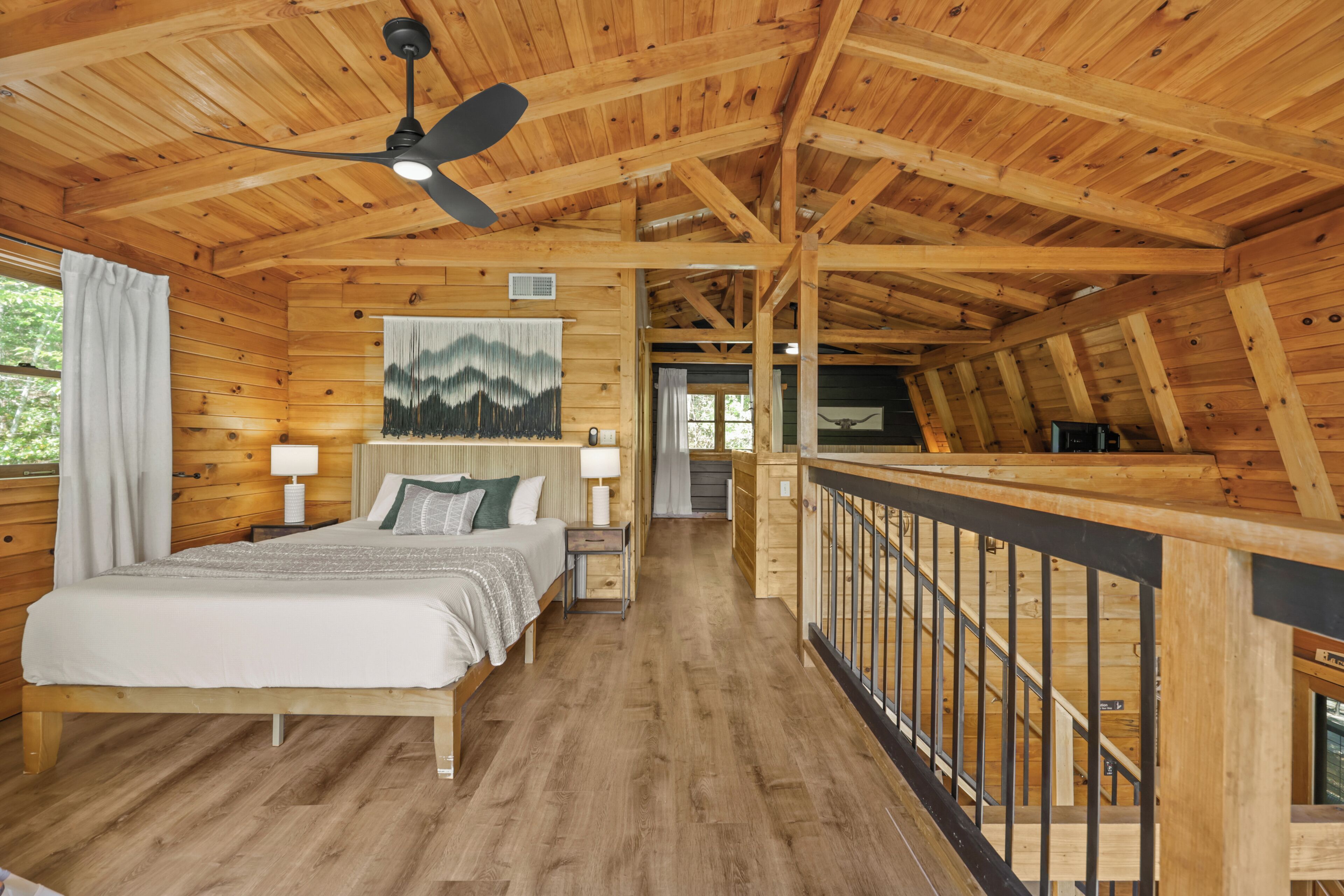 A wooden cabin bedroom with a bed, nightstands, a ceiling fan, and a view of the outdoors.