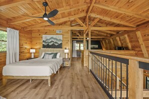 Un dormitorio de cabaña de madera con una cama, mesitas de noche, un ventilador de techo y vistas al exterior.