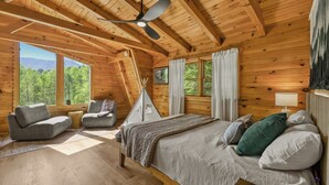 Ein Schlafzimmer in einer Blockhütte mit einem Bett, zwei Sesseln, einem kleinen Tisch, einem Deckenventilator und großen Fenstern mit Blick auf den Wald.