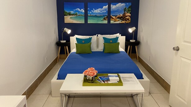 Habitación de hotel con una cama, dos mesitas de noche con lámparas, una pequeña mesa blanca con un florero y un libro, y tres cuadros en la pared.