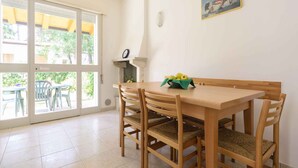 A dining area with a wooden table and chairs, a small shelf with a picture, and a view through large glass doors. - Holiday apartment with terrace (Lignano)