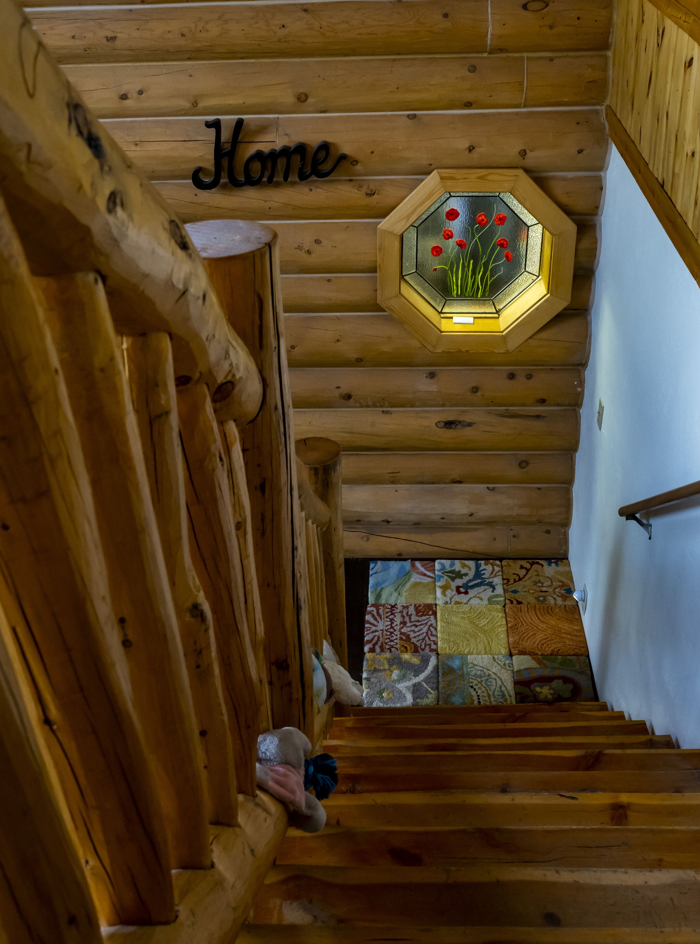 Een houten trap met een 'Home' bord erboven, een zeshoekig raam met rode bloemen, en een kleurrijke muur met een patroon aan de onderkant.