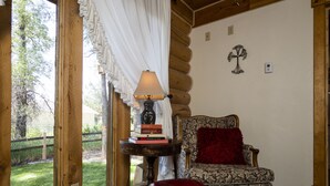 Une pièce chaleureuse avec un mur en bois brut, un fauteuil à motifs, un pouf rouge et une table sur laquelle sont posés des livres et une lampe.