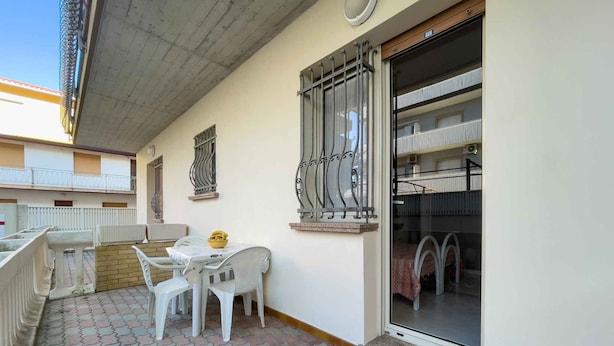 An outdoor balcony with a table and chairs, a window with decorative iron bars, and a view of the building's interior. - Holiday apartment with air conditioning (Lignano)