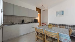 A kitchen with white cabinets, a gas stove, and a coffee maker. There is a dining table with chairs and a bowl of fruit on it. - Holiday apartment with air conditioning (Lignano)