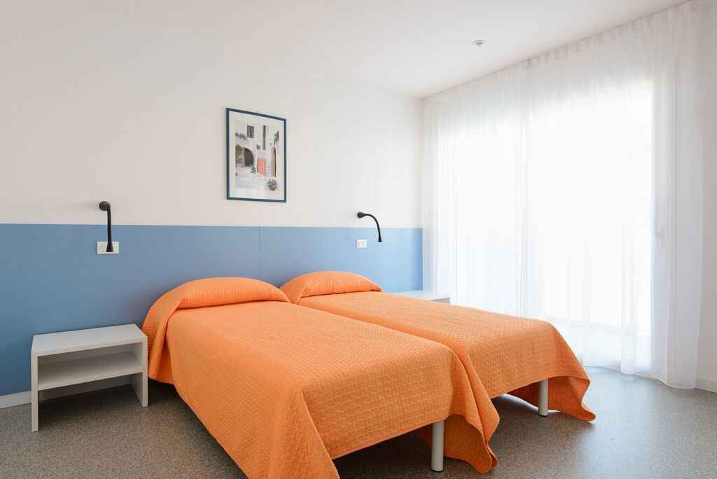 A room with two beds, a small bedside table, a wall-mounted light, and a window with curtains.