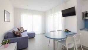 Un salon moderne comprenant un canapé, une table à manger avec des chaises, un téléviseur à écran plat fixé au mur et un coin cuisine avec un four à micro-ondes.