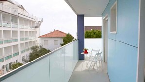Un balcon avec une balustrade en verre, une petite table surmontée d’un pot rouge et des chaises blanches.