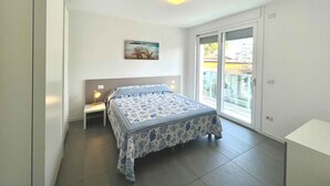 A bedroom with a bed, a nightstand, a wall-mounted light, and a window with a view of the outside. - Holiday apartment with balcony, air conditioning and internet (Lignano)