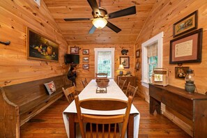 A wooden cabin interior with a dining table, chairs, a ceiling fan, and various framed pictures on the walls. - Georgia Bell's| Vintage Farmhouse by Chattahoochee (Helen)