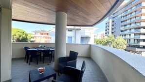 A balcony with wicker furniture, a table with a bowl, and a view of surrounding buildings. - Holiday apartment with balcony and air conditioning (Lignano)