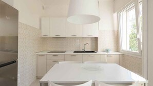 A modern kitchen with white cabinets, a stainless steel refrigerator, a white dining table with two chairs, and a window providing natural light. - Holiday apartment mit Klimaanlage und Balkon (Lignano)