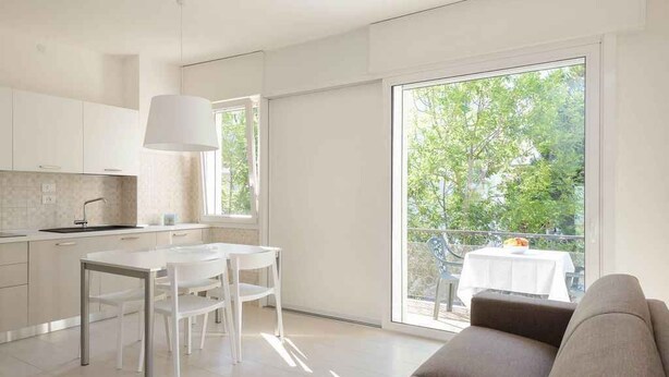 A modern kitchen with white cabinets and a dining area with a white table and chairs. There is a large sliding glass door leading to a balcony with a table and chairs. - Holiday apartment mit Klimaanlage und Balkon (Lignano)