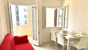 Un appartement moderne avec un canapé rouge, un coin repas avec des chaises blanches et un balcon offrant une vue sur les immeubles.