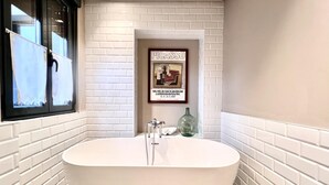 Une salle de bain avec une baignoire sur pieds, des murs recouverts de carreaux blancs, un tableau encadré au mur et une fenêtre avec des rideaux.