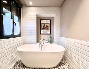 A bathroom with a freestanding bathtub, white tiled walls, a framed picture on the wall, and a window with curtains.