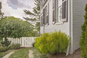 A white brick house with a white picket fence, a small garden with green shrubs, and a pathway leading to the house.