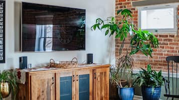 Een kamer met een bakstenen muur, een houten kast en een groot schilderij. Er staan potplanten en er is een raam.