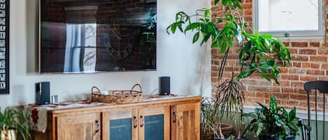 Une pièce avec un mur en briques, un meuble en bois et un grand tableau. On y trouve des plantes en pot et une fenêtre.