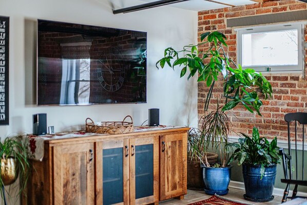 Una habitación con pared de ladrillo, un armario de madera y un cuadro grande. Hay plantas en macetas y una ventana.