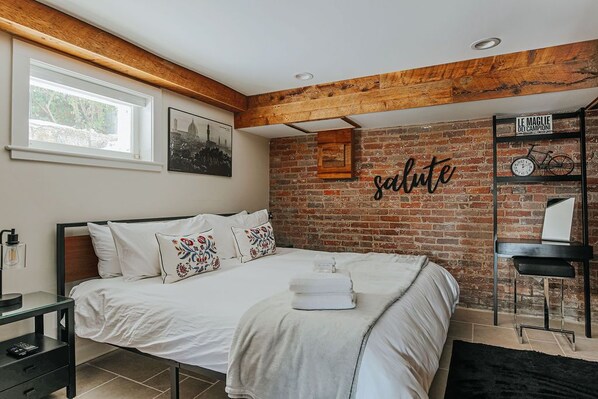 A bedroom with a brick wall, a bed with white bedding, a wooden ceiling, and a shelf with a bicycle and a sign.