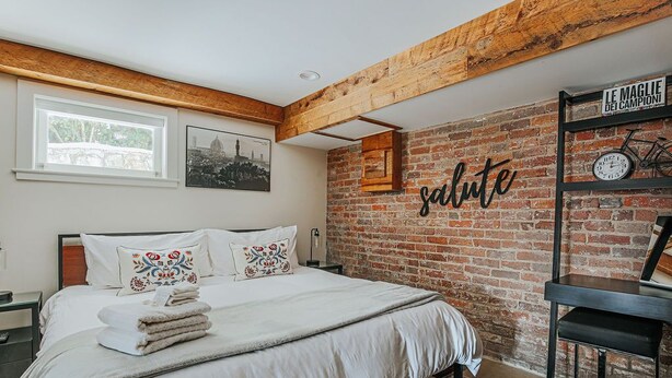 A bedroom with a brick wall, a bed with white bedding, a nightstand, a window, and a wall-mounted shelf with a clock and a bicycle.