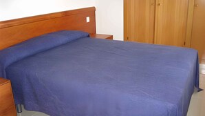 A bedroom with a wooden bed frame, a blue bedspread, a wooden nightstand, and a wardrobe with two doors. - Holiday apartment with pool (Roses)