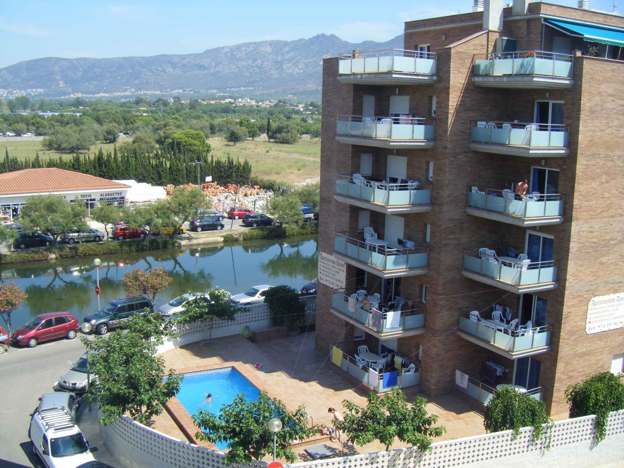 Un immeuble d’appartements à plusieurs étages, doté de balcons, d’une piscine et offrant une vue sur un lac et des montagnes en arrière-plan.