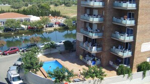 A multi-story apartment building with balconies, a swimming pool, and a view of a lake and mountains in the background. - Holiday apartment with pool (Roses)