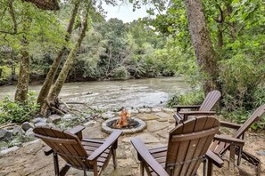 Una chimenea rodeada de sillas Adirondack en un área boscosa con un río.