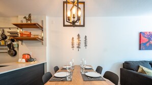A modern dining area with a wooden table set for four, surrounded by black chairs. Above the table, there is a wall-mounted light fixture with three bulbs. The wall features three black tree silhouettes.