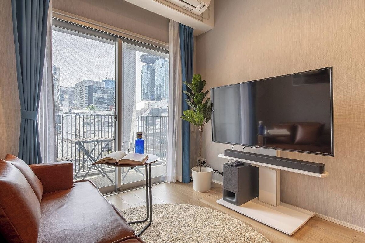 A modern living room with a large flat-screen TV, a leather sofa, a small table with a book, and a potted plant.