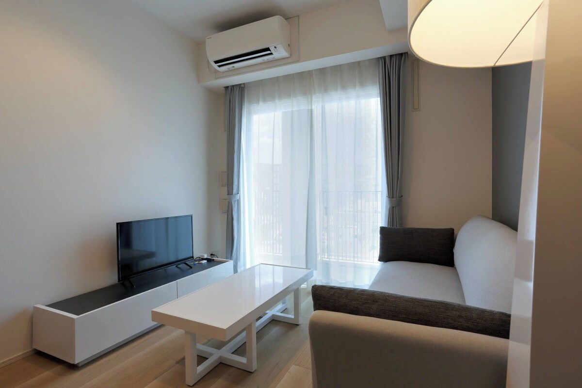 A modern hotel room with a flat-screen TV, a white table, a sofa, and a large window with curtains.