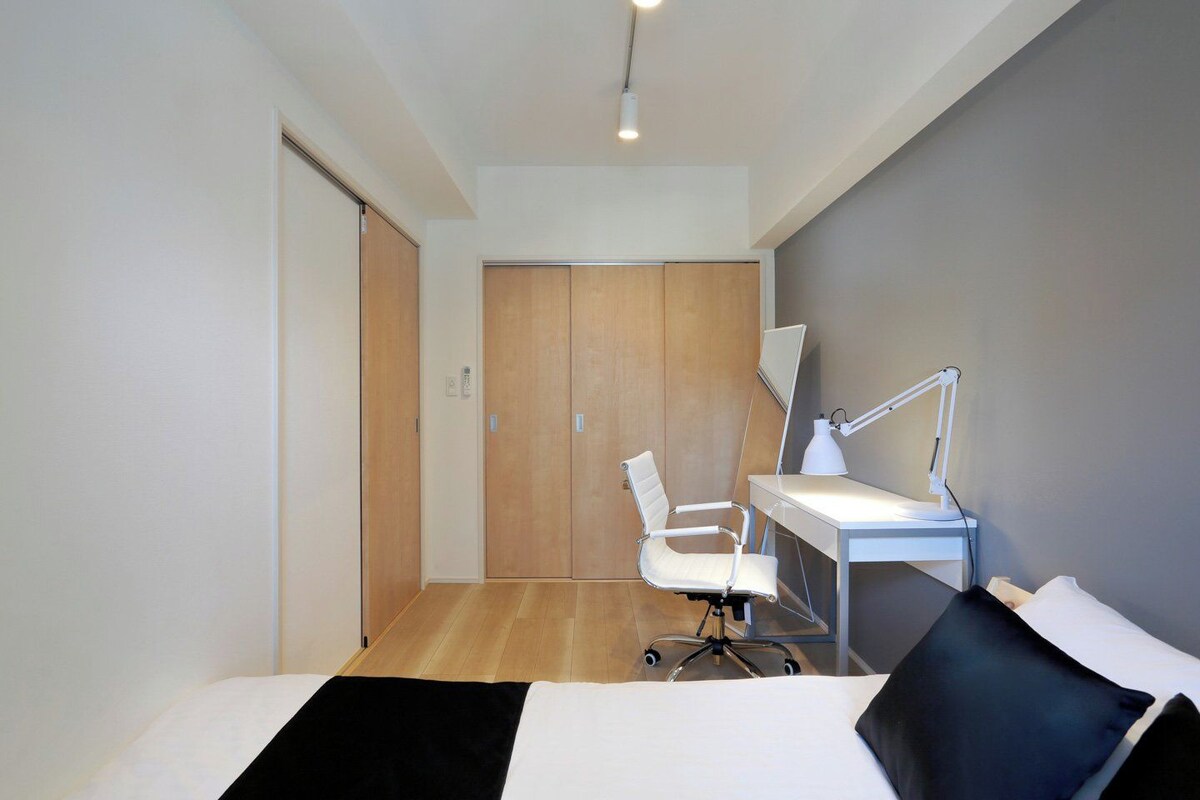 A hotel room with a bed, a desk with a chair, a desk lamp, and a wooden wardrobe.