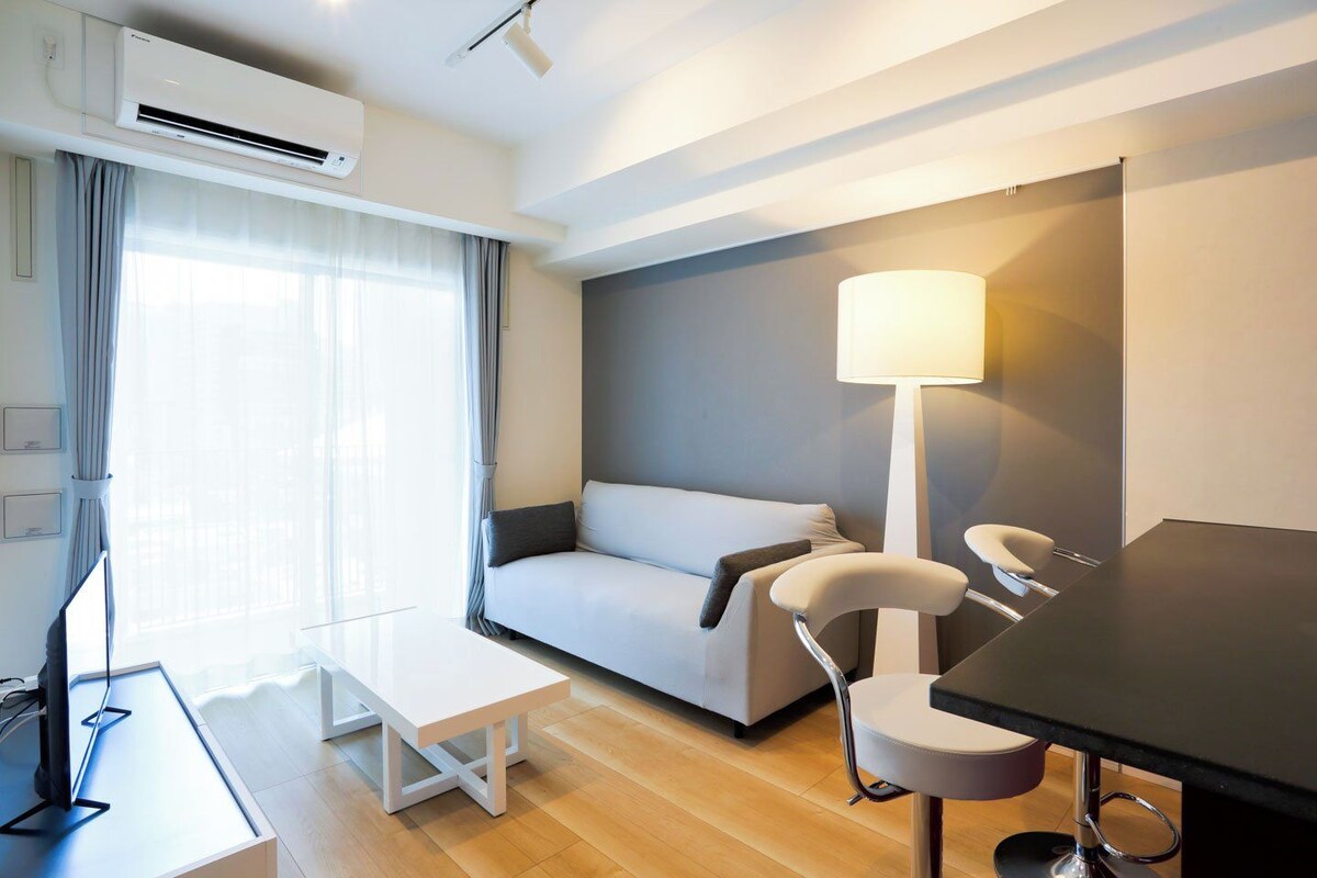 A modern hotel room with a sofa, a desk with a computer, a lamp, and an air conditioning unit.