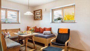 A cozy room with a wooden dining table, a sofa, a chair, and a bed. There are windows with views of trees and a shelf with decorative items.